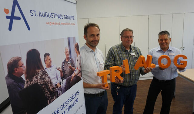 Drei Männer stehen neben einem Aufsteller der Augustinus Gruppe. Die drei Männer halten Buchstaben in der Hand, die zusammen das Wort Trialog bilden. 