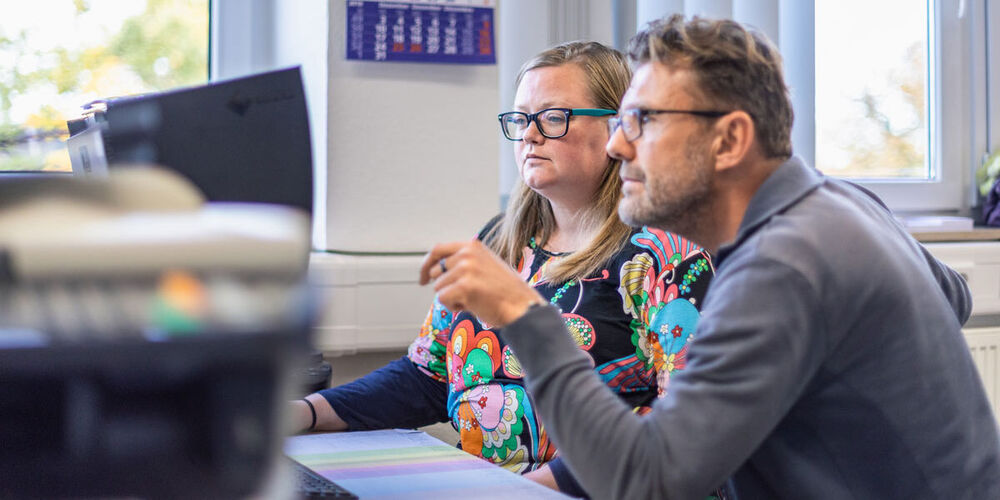 Ein Mann und eine Frau sitzen gemeinsam an einem Schreibtisch und schauen auf einen Computerbildschirm. 