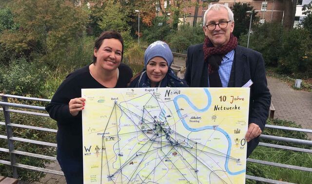 Zwei Frauen und ein Mann stehen auf einer Terrasse und halten ein großes Plakat in der Hand auf dem die unterschiedlichen Verbindungslinien des Netzwerkes der Behindertenhilfe abgebildet sind. 