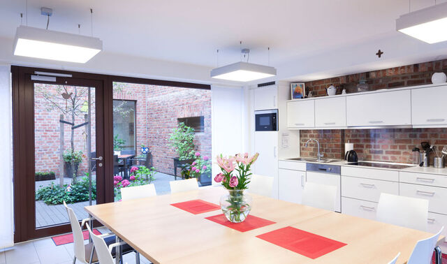 Große moderne Küche mit einem großen Tisch und vielen Stühlen. Im Hintergrund ist eine große Glasfront, welche einen Innenhof zeigt. 